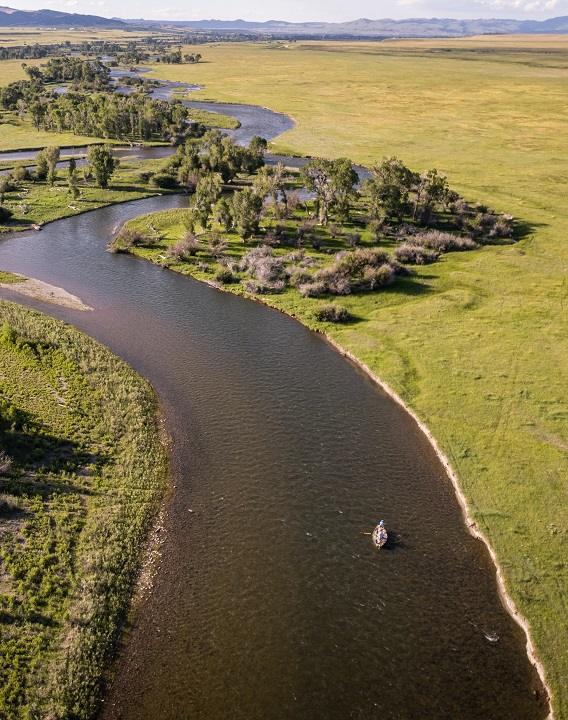 A fly fishing boat floats down a winding Montana river surrounded by green grass during a trip with The Tackle Shop.