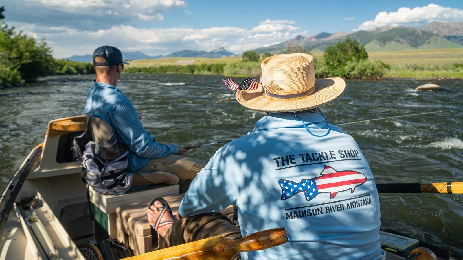 A man sits on a boat while fly fishing as a guide wearing a Tackle Shop sweatshirt steers them down a Montana river.