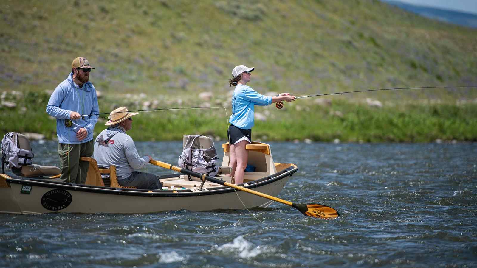 A Tackle Shop guide steers a boat down a river while a man and a woman fly fish during a summer trip.