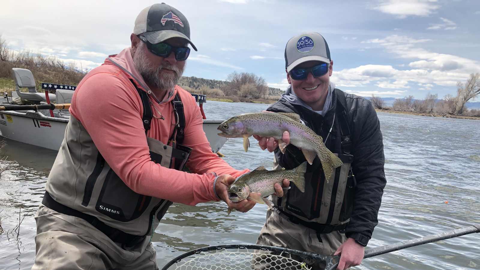 Two men hold fish while standing in a Montana river as a boat floats in the background during a trip with The Tackle Shop.