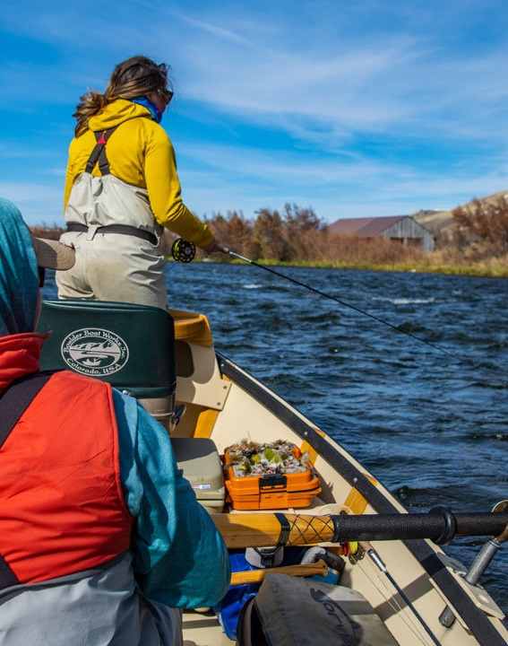 A guide steers a boat downriver while a woman stands and fly fishes during a trip with The Tackle Shop.