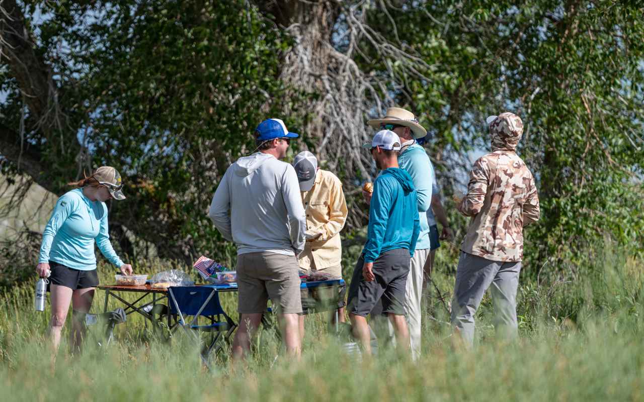 A group of people stand in a grassy area surrounding a table with lunch sandwiches during a trip with The Tackle Shop.